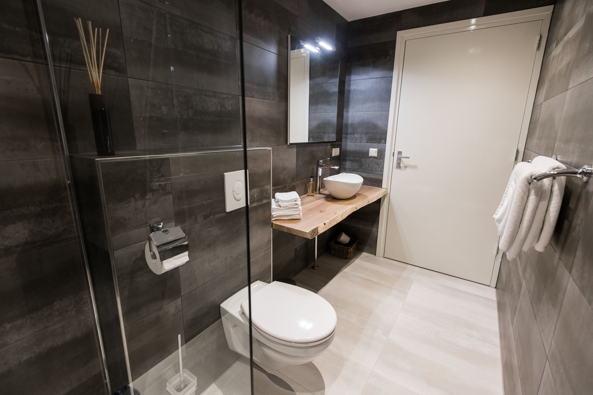 Modern bathroom with a shower area and bath tub including a wall mirror beside a fancy plant near a tap and sink over the wooden counter and dark cupboard